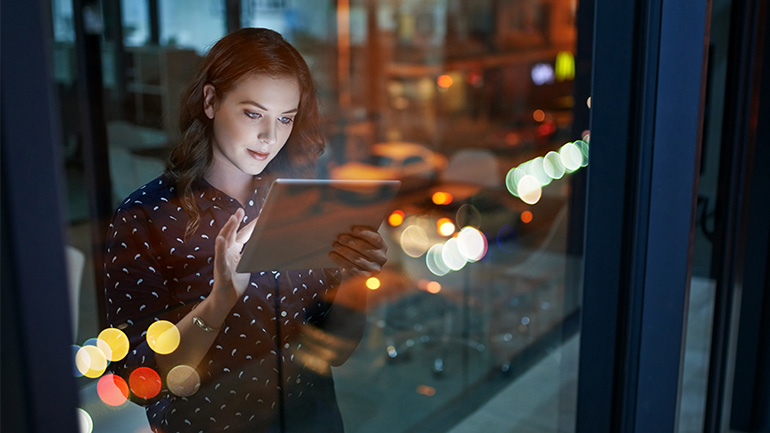 A person using a tablet device near a window at night, with city lights and reflections visible outside