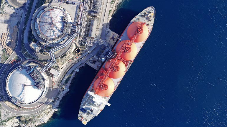 An aerial view of a large liquefied natural gas carrier ship docked at a port with several large storage tanks on land and connected by pipes to the ship.