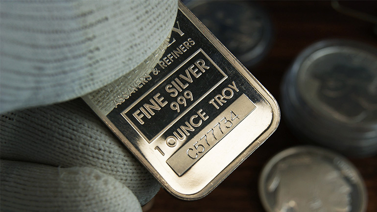 Fine silver bar held in gloved hand, with coins in the background.