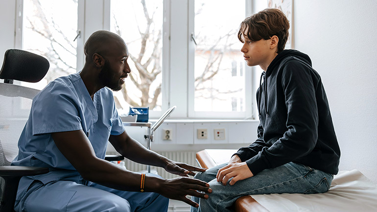 Clinician talking with a young patient seated on an exam table in a medical room.