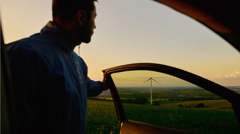Person looking across a countryside wind farm through an open car door at sunset.