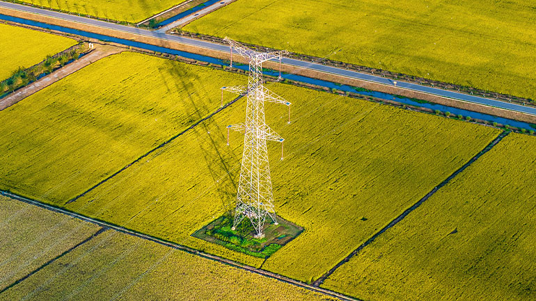 Electrical transmission tower standing in a yellow agricultural field, viewed from above, with nearby road and irrigation canal.