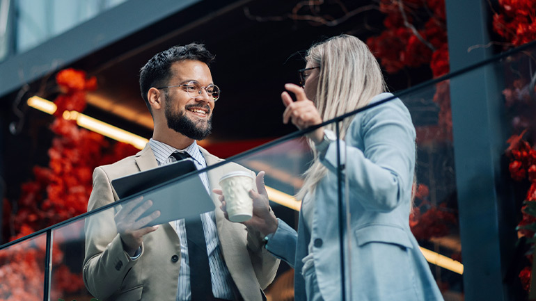 Two professionals talking on a modern office balcony, holding a tablet and coffee.