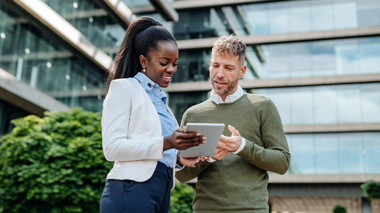 Two people standing outside a modern glass building, looking at a tablet computer together.