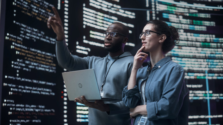 Two people analyzing data on a wall while holding a laptop computer.