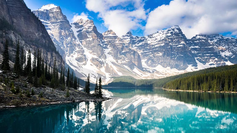Wide landscape view of the Canadian Rocky Mountains with a blue lake in the foreground.