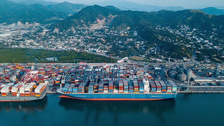 Aerial view of a container ship docked at a busy port with stacked shipping containers and a hillside city in the background.