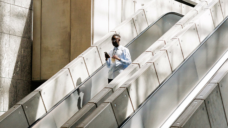 Person using a smartphone while descending a modern indoor escalator with sunlight casting shadows across the structure.