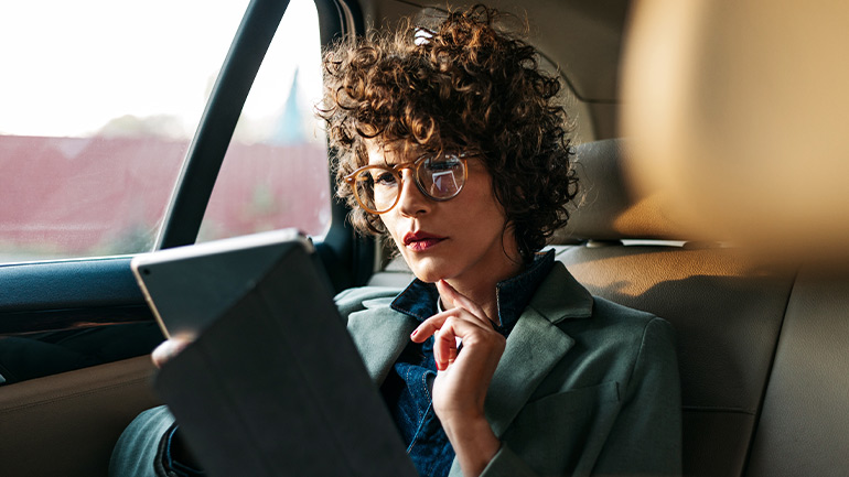 Person sitting in a car’s back seat, using a tablet with one hand and wearing glasses.
