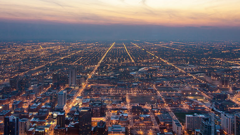 Aerial view of a city at dusk, with illuminated streets stretching to the horizon beneath a colorful sunset sky.