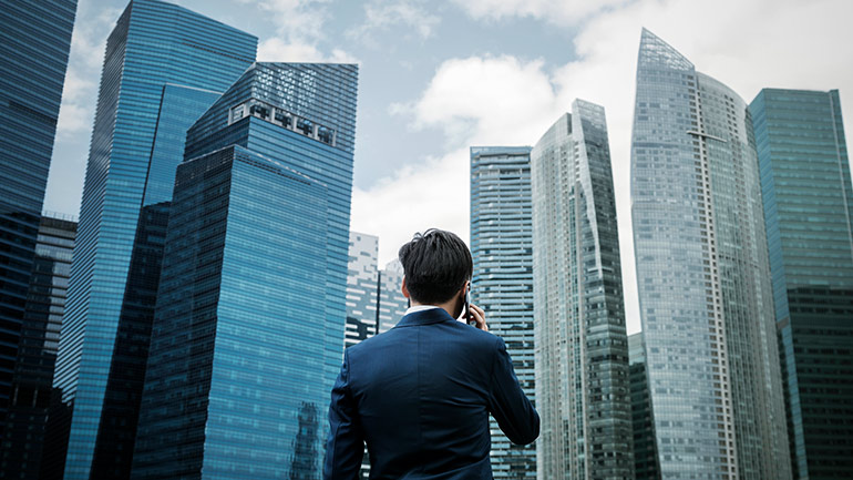 A man stands in front of multiple city skyscrapers while holding a cellphone to his ear.