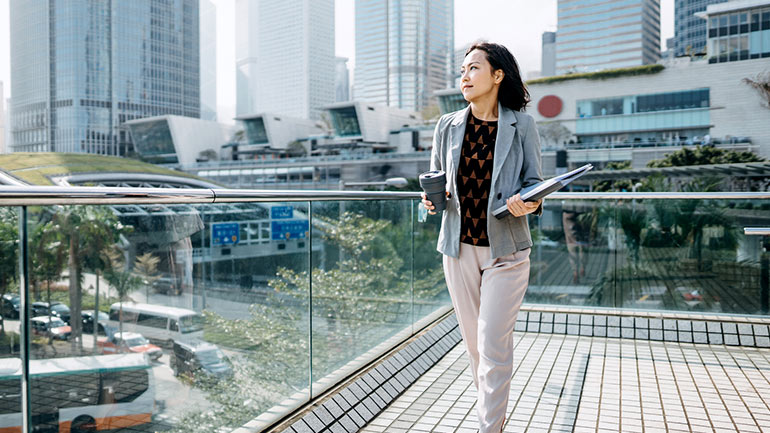 Business professional walking on an urban terrace with a coffee and documents, city skyline in the background.