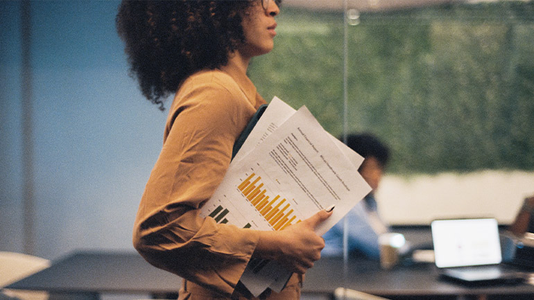 Worker carrying printed reports through a meeting room with colleagues and laptops in the background.