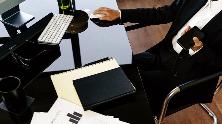Professional seated at desk using computer mouse, with smartphone, notebook and keyboard on workspace.