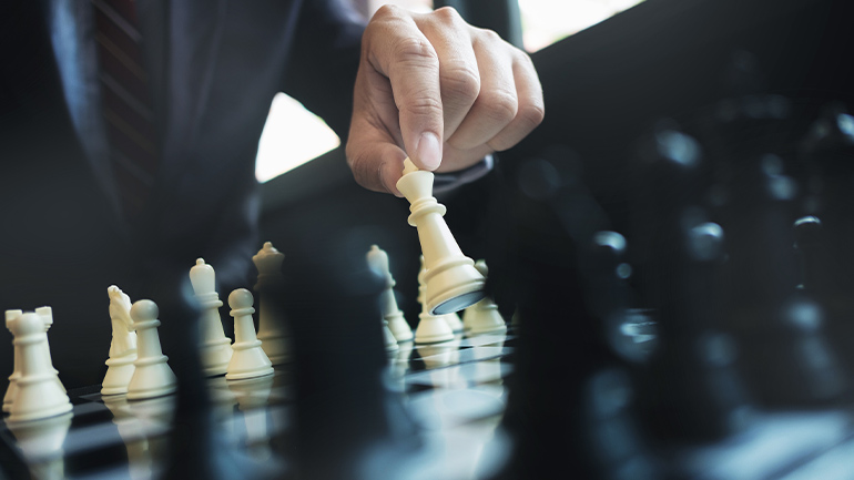 Close-up of a business professional's hand moving a white chess piece on a chessboard, with other pieces in the foreground and background.