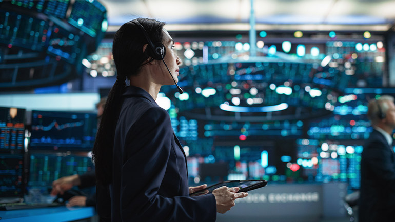 Person holding a tablet in a financial trading room with digital screens displaying market data.