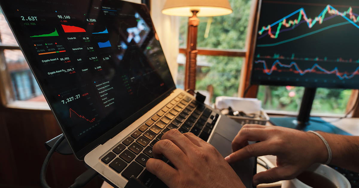Person using a laptop displaying market data and asset prices beside a monitor showing financial charts and trading trends.