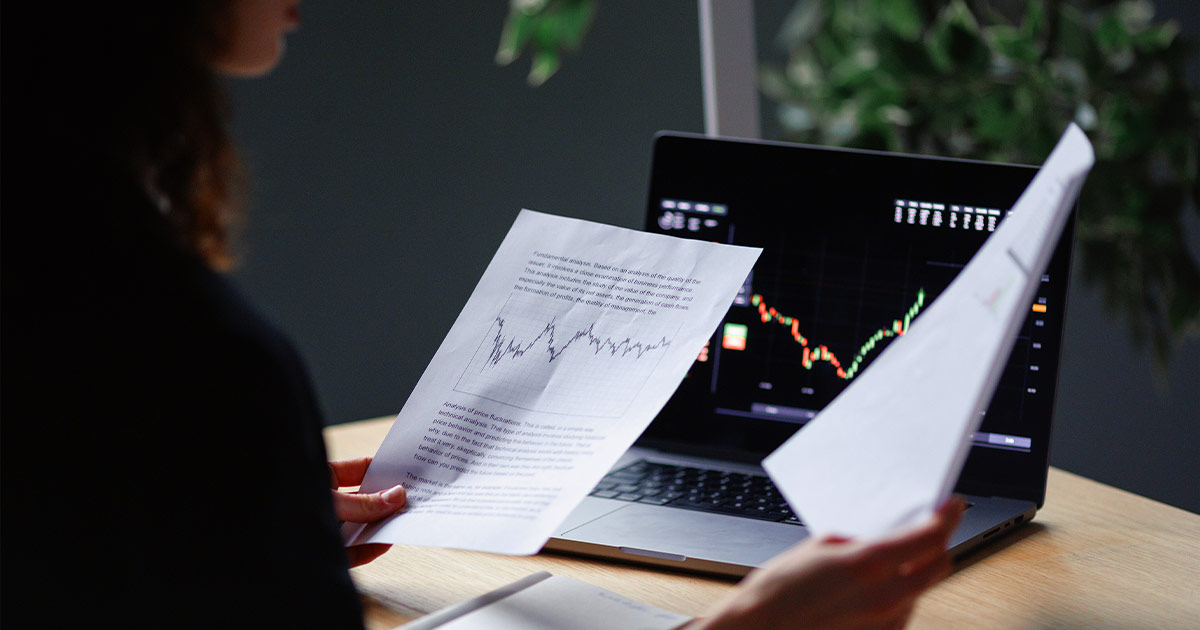 Person holding printed financial charts in front of a laptop showing a market graph.