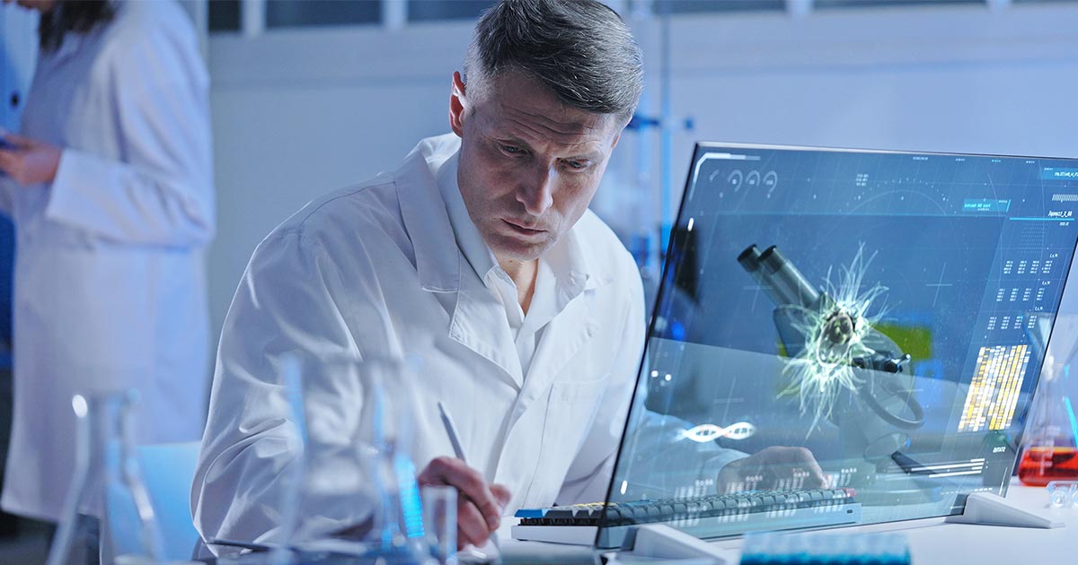 Scientist in a lab coat working at a computer with a digital microscope interface and laboratory glassware.