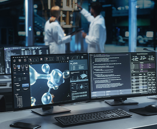 A modern laboratory workspace featuring two large computer monitors on a desk, displaying scientific data. Two people in lab coats are in the background discussing something near scientific equipment.